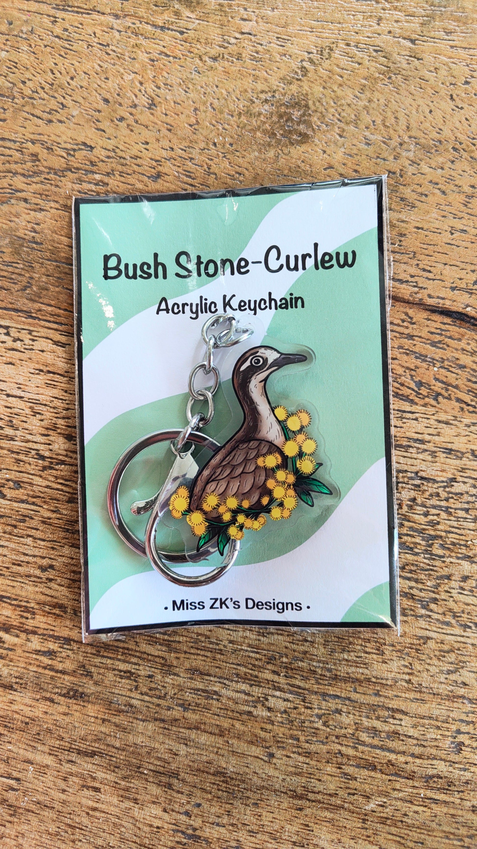 An acrylic keychain featuring a curlew bird and yellow wattle flowers, presented on its original packaging card which reads 'Bush Stone-Curlew Acrylic Keychain' by 'Miss ZK's Designs', set on a wooden surface.