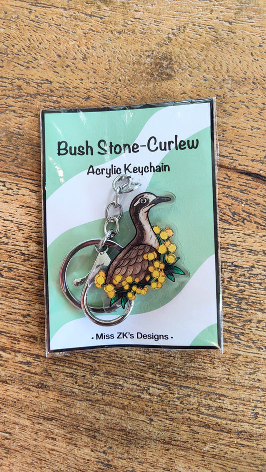 An acrylic keychain featuring a curlew bird and yellow wattle flowers, presented on its original packaging card which reads 'Bush Stone-Curlew Acrylic Keychain' by 'Miss ZK's Designs', set on a wooden surface.